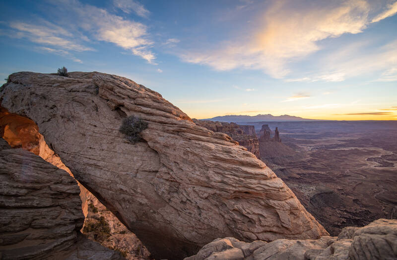 Photo of Mesa Arch from an alternate perspective showing mountains and mesas in the background.