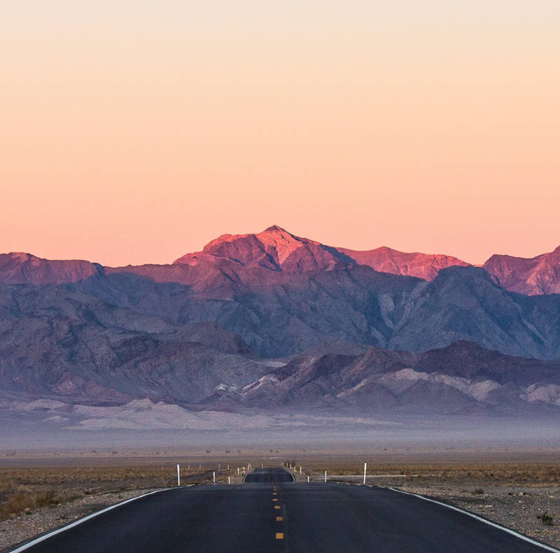 Photo of a road at sunrise and mountains with alpenglow in the background.